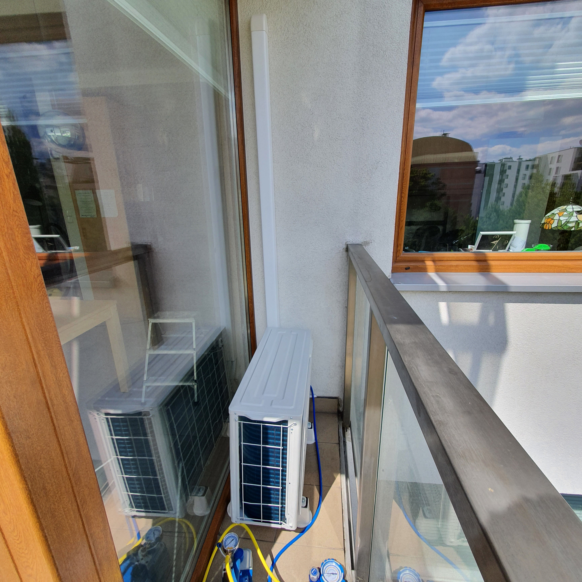 Biała jednostka zewnętrzna klimatyzacji na balkonie, podłączona do manometrów za pomocą niebieskich i żółtych przewodów, widoczna przez szybę odbita druga jednostka.