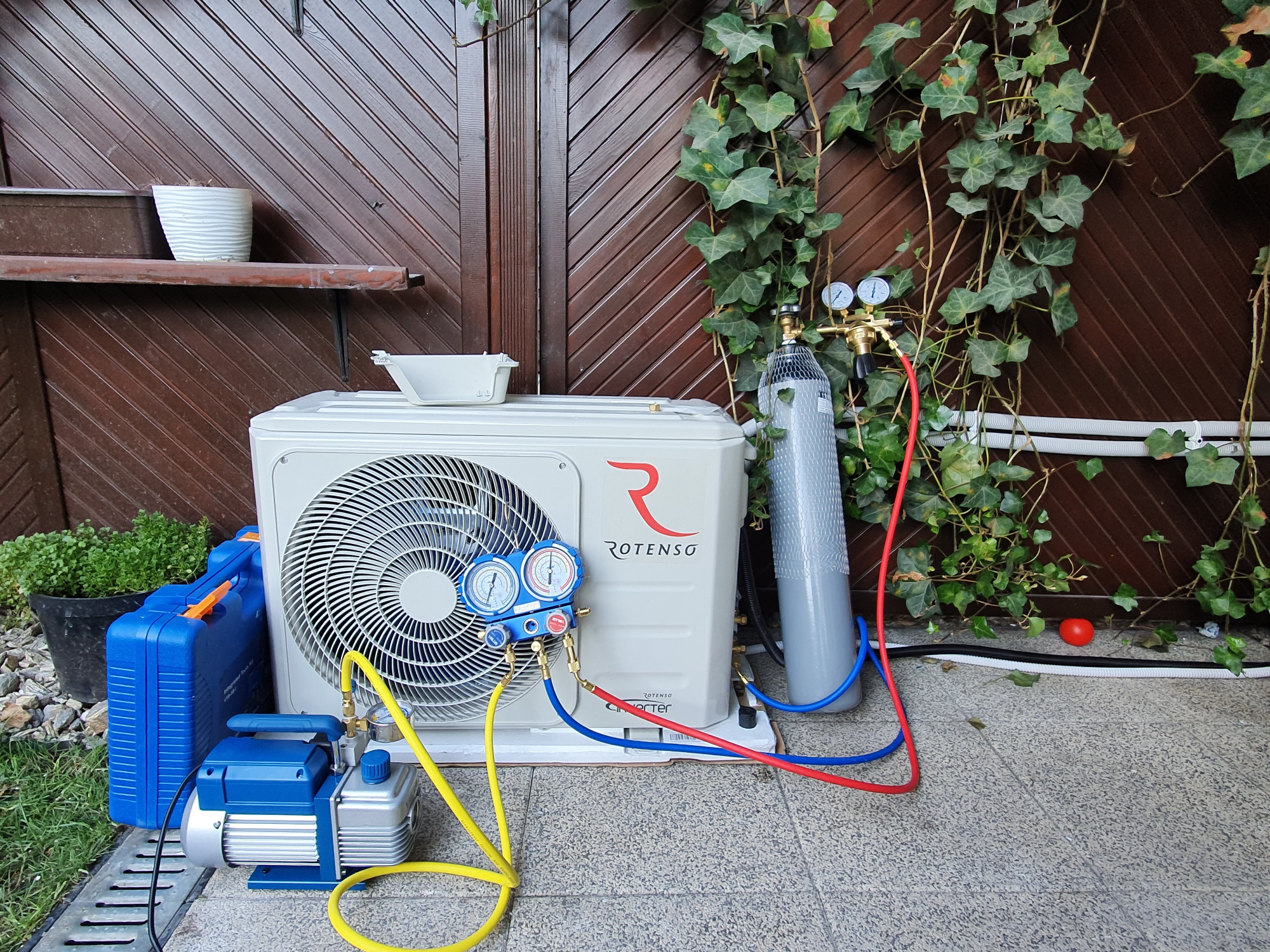 Serwis klimatyzacji Rotenso w Poznaniu: Manometr podłączony do jednostki zewnętrznej, obok butla z gazem i pompa próżniowa, na tle brązowej drewnianej ściany z bluszczem.