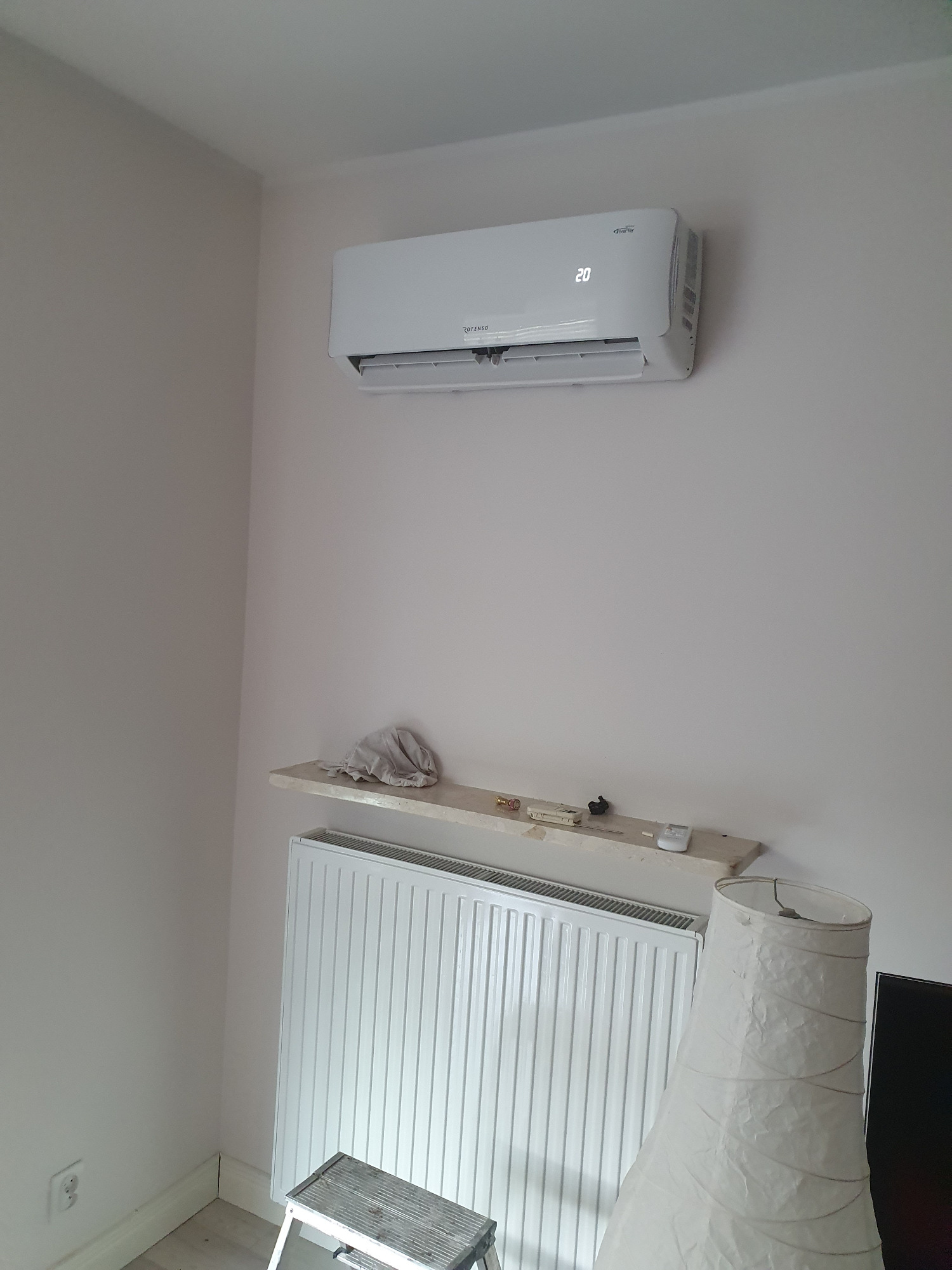 Biały klimatyzator Rotenso z wyświetlaczem temperatury 20 stopni Celsjusza zamontowany na jasnej ścianie w pokoju, pod nim półka z narzędziami i biały grzejnik panelowy.