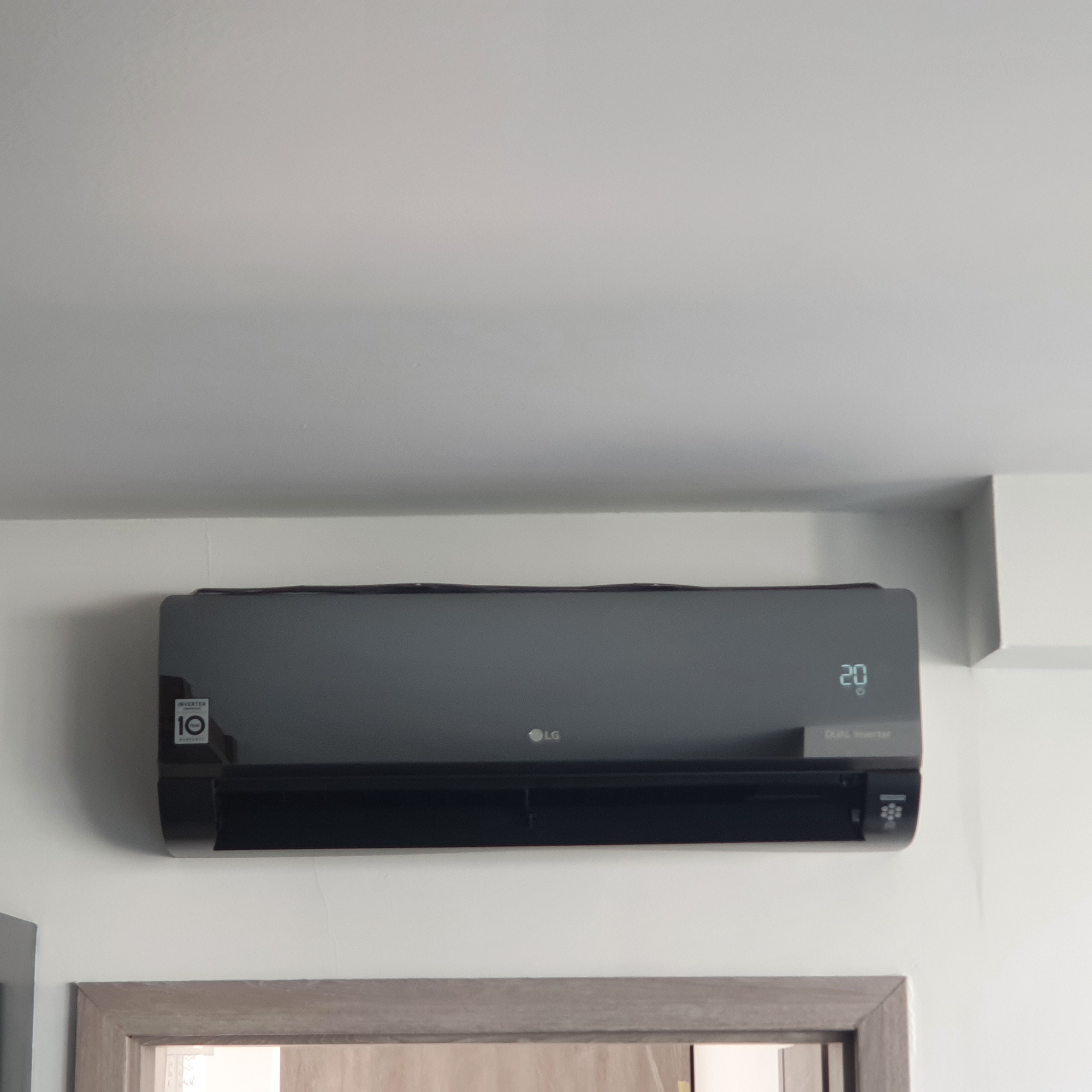 Czarny klimatyzator LG Dual Inverter zamontowany na białej ścianie, wyświetlający temperaturę 20 stopni Celsjusza, widoczny fragment futryny drzwi na dole kadru.