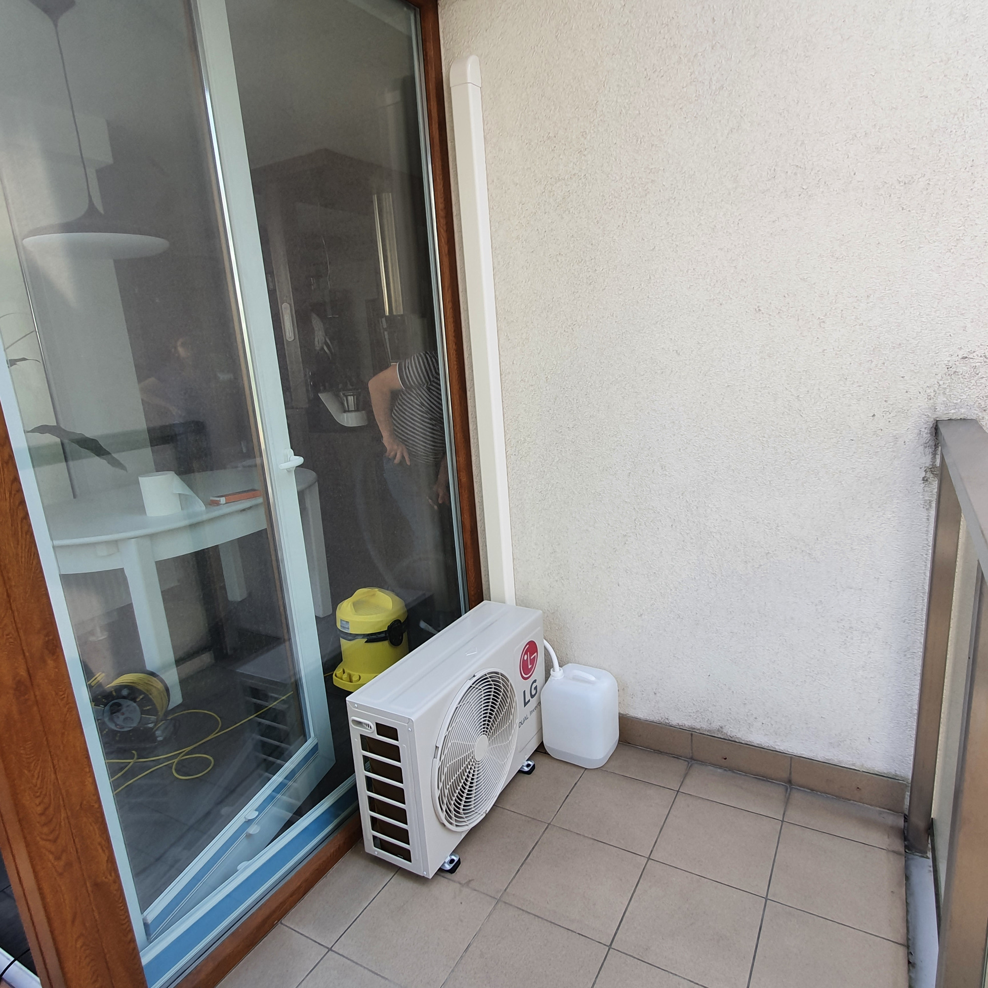 Biała jednostka zewnętrzna klimatyzacji LG Dual Inverter, umieszczona na balkonie obok okna balkonowego, obok niej biały kanister, w tle widoczny odkurzacz i osoba w paski.