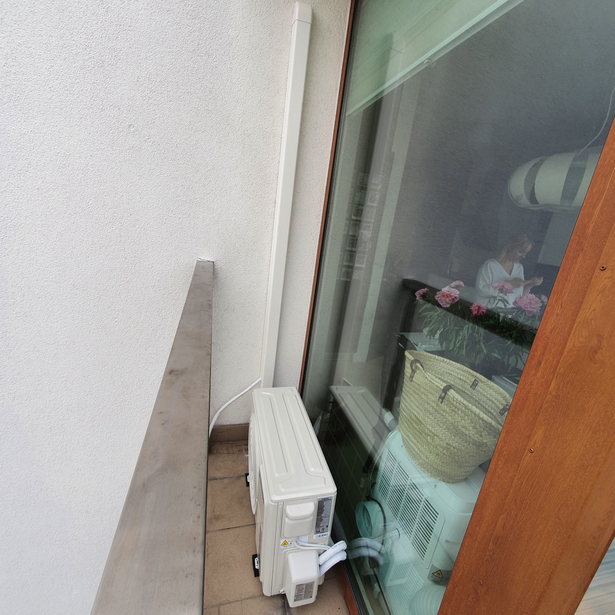 Przenośny klimatyzator ustawiony na balkonie, widoczny przez szybę, z elementami wnętrza odbijającymi się w szkle.