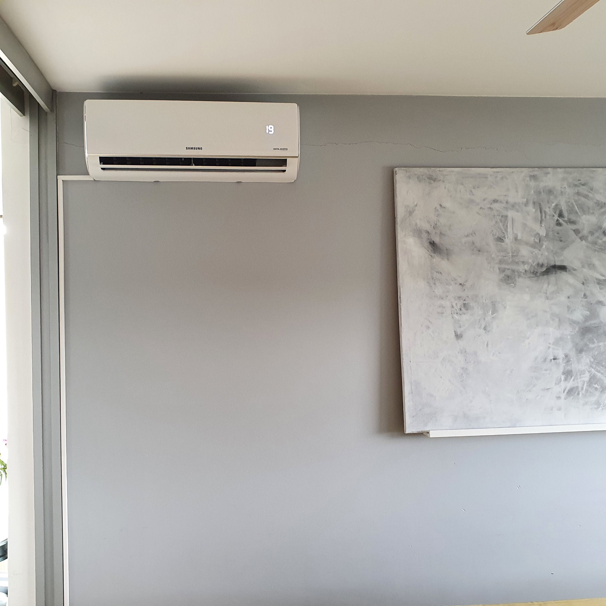 Biała klimatyzacja marki Samsung zamontowana na szarej ścianie w pomieszczeniu z abstrakcyjnym obrazem w tle, ustawiona na temperaturę 19 stopni Celsjusza.