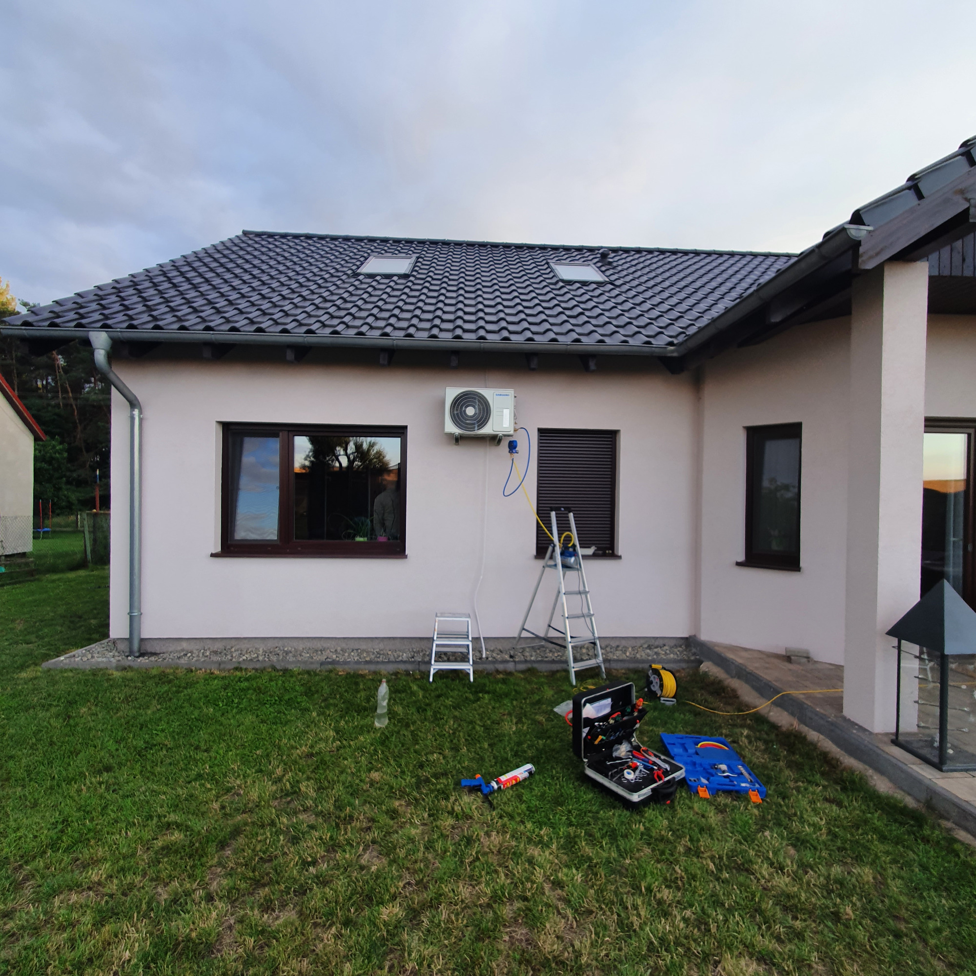 Montaż klimatyzatora na elewacji domu jednorodzinnego, widoczne narzędzia, drabiny i jednostka zewnętrzna, trawnik i fragment elewacji.