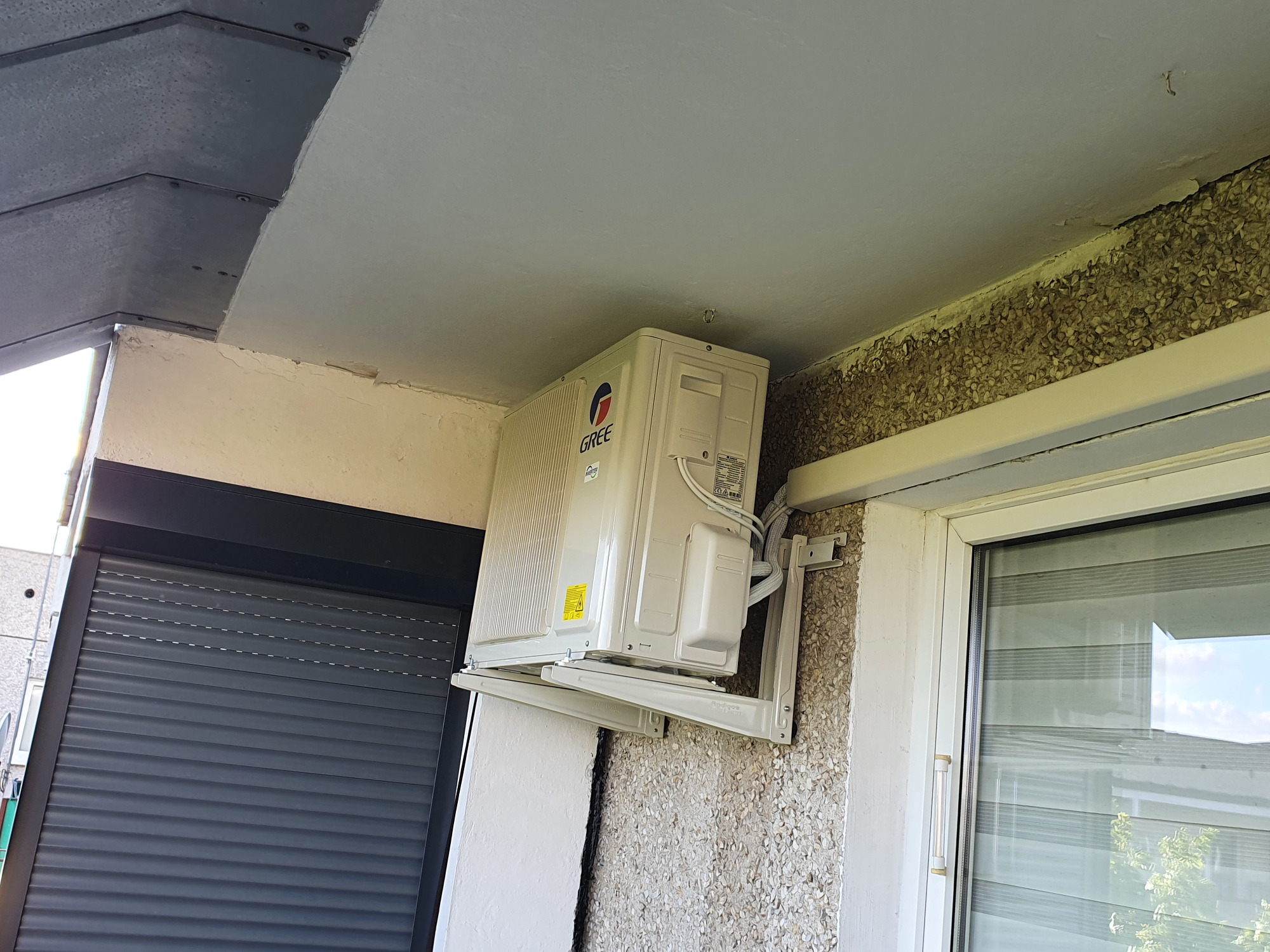 Zewnętrzna jednostka klimatyzatora marki Gree zamontowana na balkonie bloku mieszkalnego, widoczne okno i roleta.