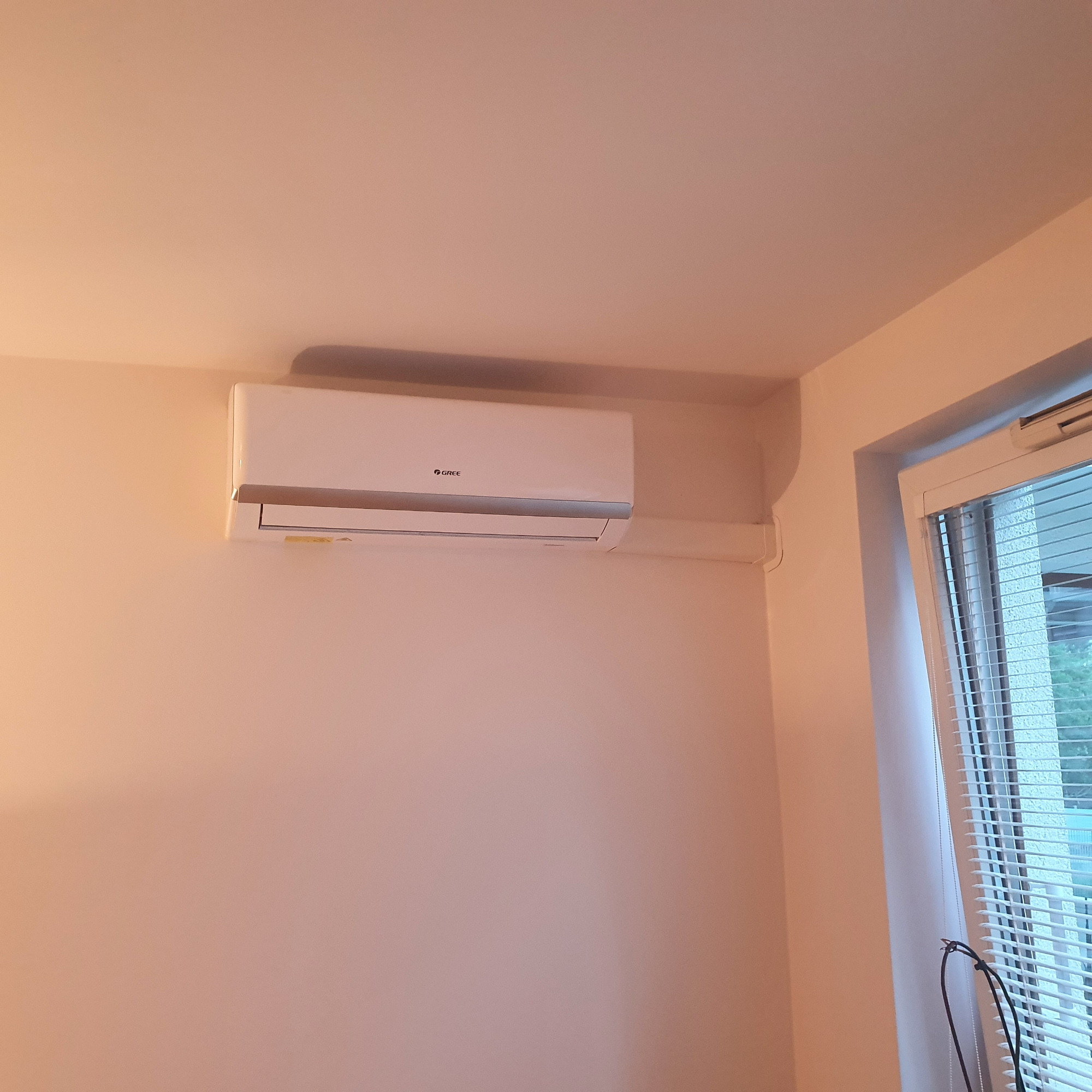 Biały klimatyzator marki Gree zamontowany na ścianie w jasnym pomieszczeniu, widoczny fragment okna z żaluzjami.