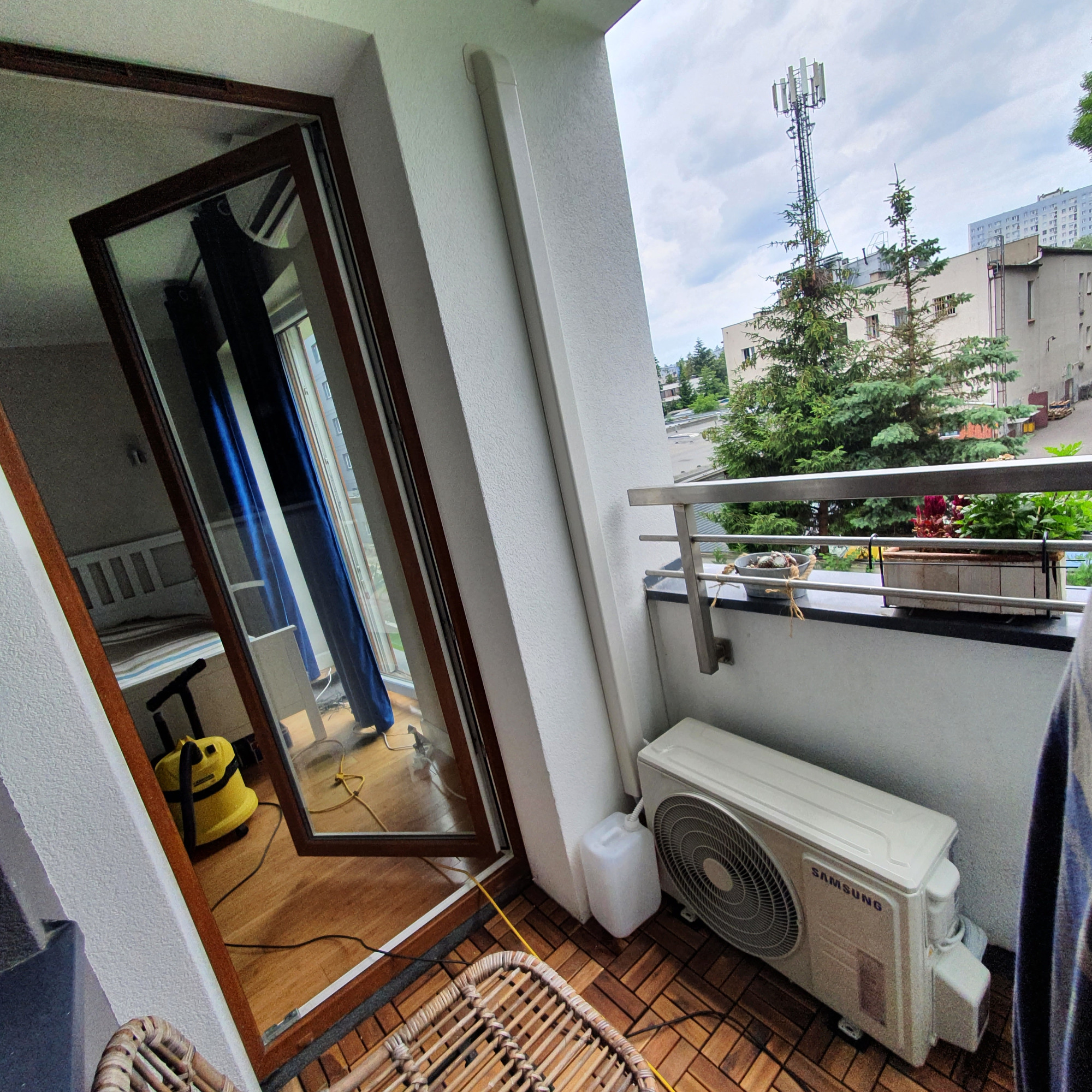 Biała jednostka zewnętrzna klimatyzacji Samsung zamontowana na balkonie z drewnianą podłogą, widok na zewnątrz z wieżą telekomunikacyjną w tle, w odbiciu w otwartym oknie widać odkurzacz i łóżko.
