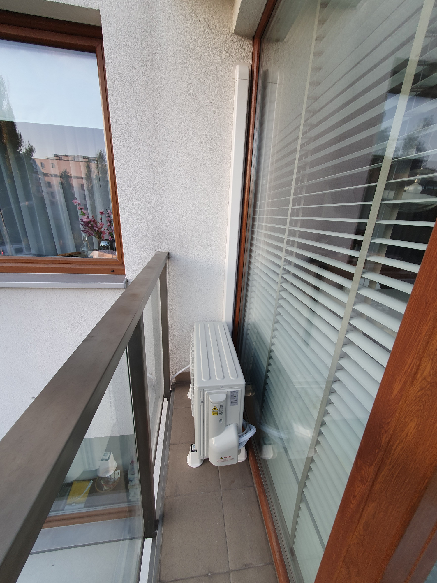 Zewnętrzna jednostka klimatyzatora zamontowana na balkonie z widoczną balustradą i oknem z żaluzjami.