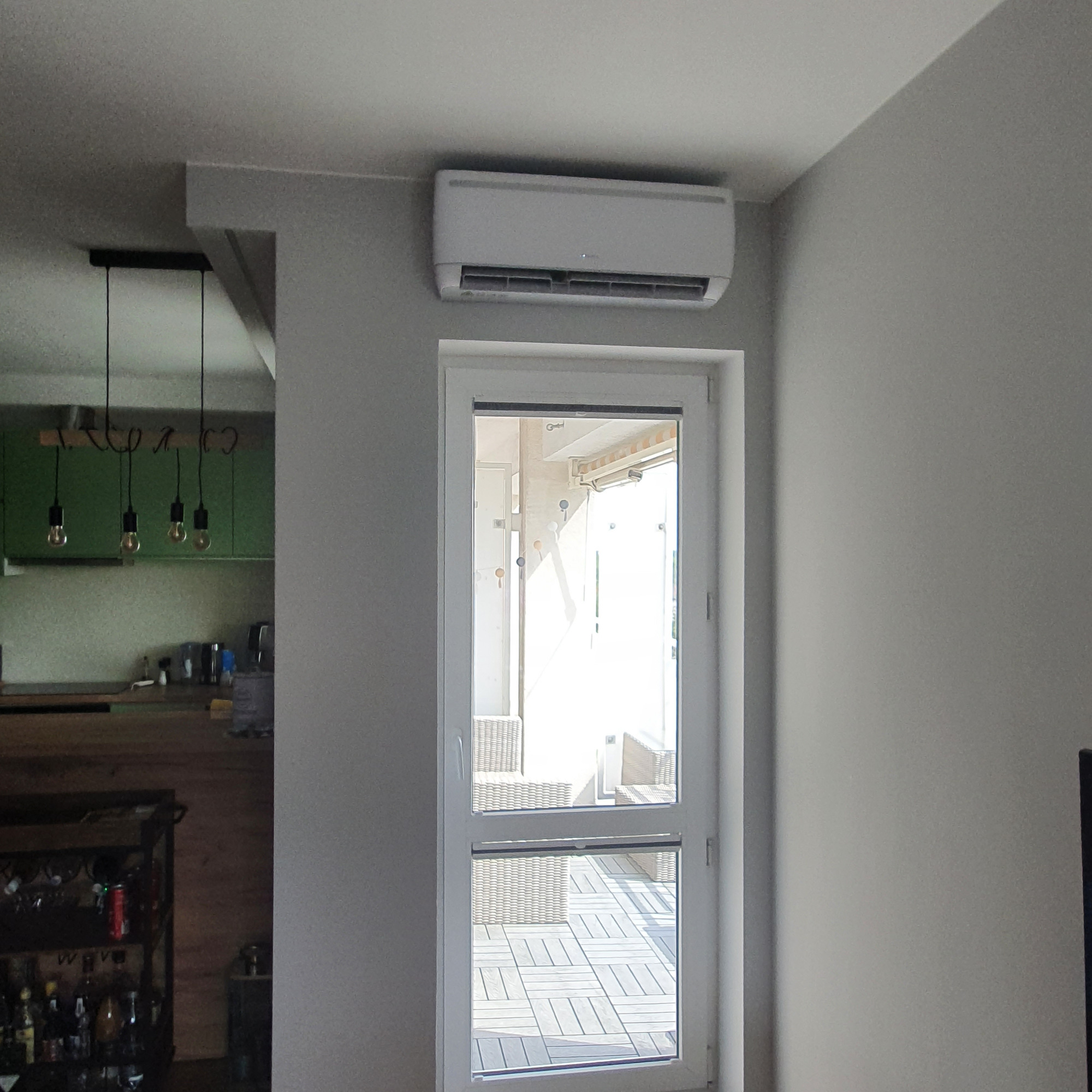 Biała jednostka klimatyzacji zamontowana nad balkonowymi drzwiami w jasnym wnętrzu domu, widoczna część kuchni z wiszącymi lampami.