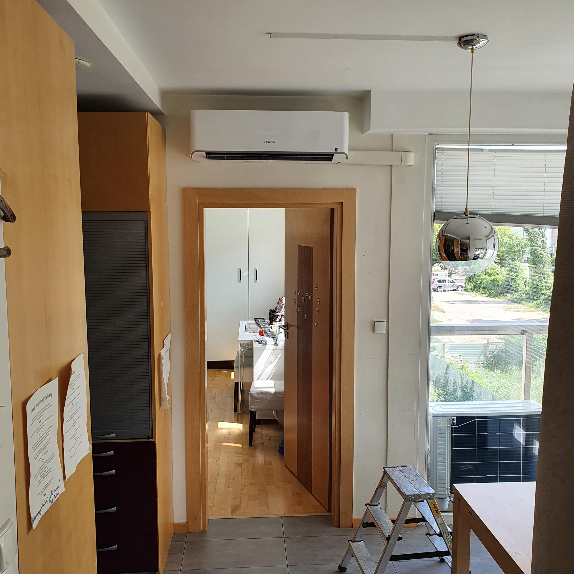 Wnętrze mieszkania z zamontowanym klimatyzatorem Hisense na ścianie nad drzwiami, widok na pokój przez otwarte drzwi, drabina oparta o ścianę w pobliżu okna z widoczną jednostką zewnętrzną...
