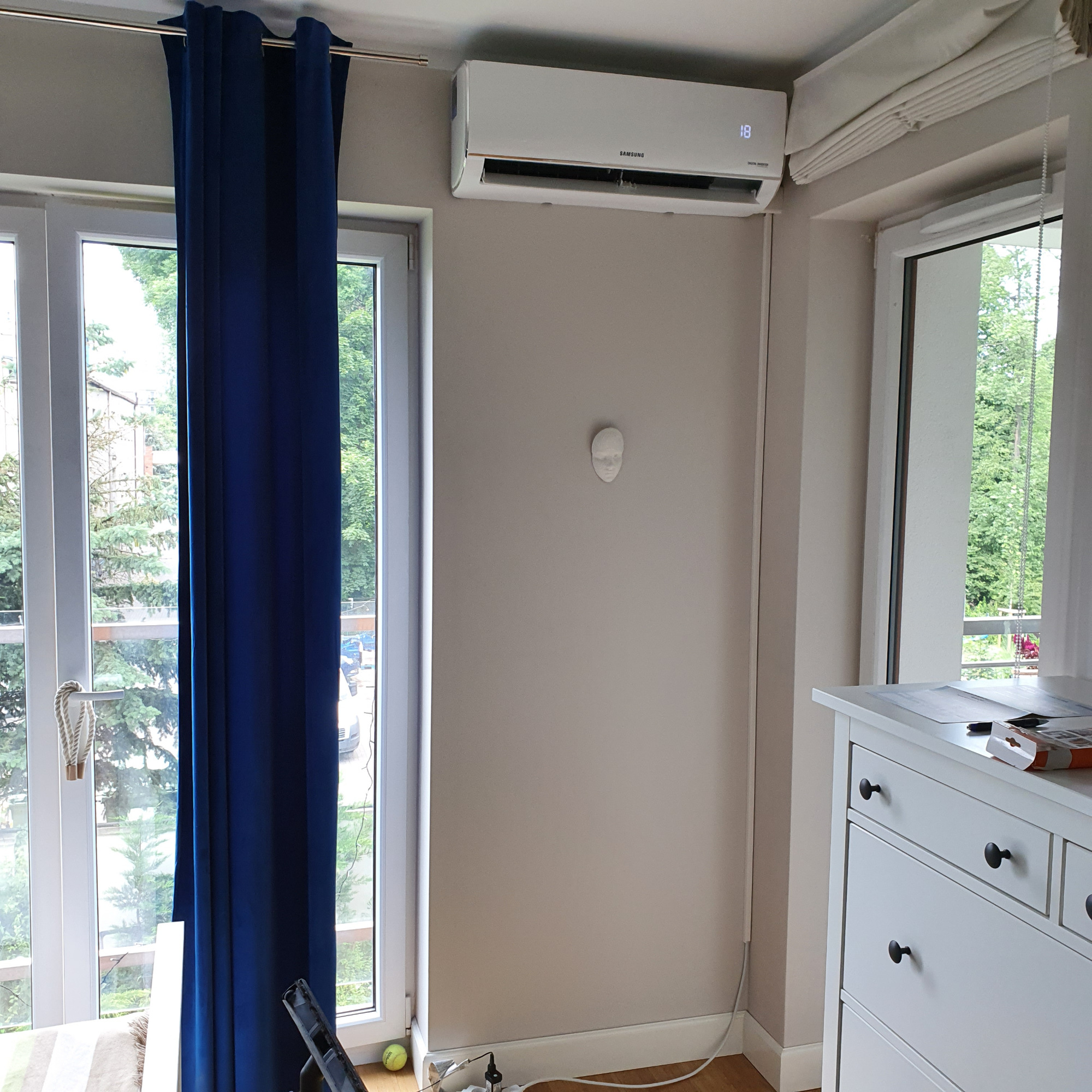 Biały klimatyzator Samsung zamontowany na ścianie w pokoju z niebieskimi zasłonami i białymi meblami, widok z dołu.