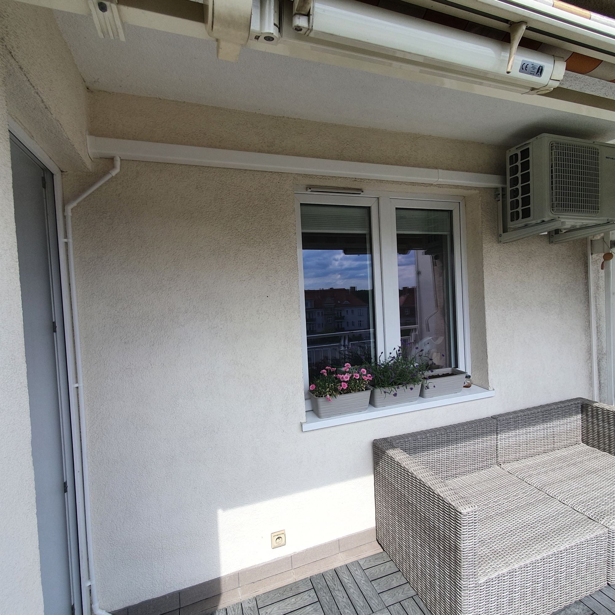 Zewnętrzna jednostka klimatyzacji zamontowana na balkonie budynku mieszkalnego, widoczne kwiaty w doniczkach na parapecie i meble ogrodowe z rattanu.