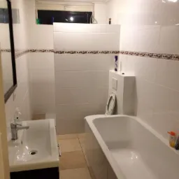 Wnętrze łazienki z białą wanną, umywalką z szafką w kolorze ciemnego brązu, toaletą ze spłuczką w zabudowie, wykończonej białymi płytkami z dekoracyjnym pasem na ścianach i brązową podłogą.