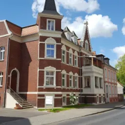 Odrestaurowana kamienica z czerwonej cegły z białymi detalami architektonicznymi, widoczna z poziomu ulicy w słoneczny dzień, z fragmentem znaku drogowego po prawej stronie.