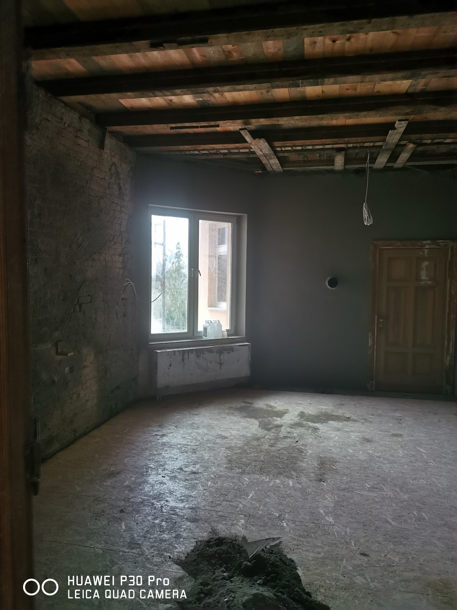 Wnętrze pokoju w trakcie prac przygotowawczych: ściana z surowej cegły, częściowo otynkowana ściana, sufit z drewnianych belek, surowa podłoga z płyt OSB, okno z grzejnikiem, drewniane drzwi...