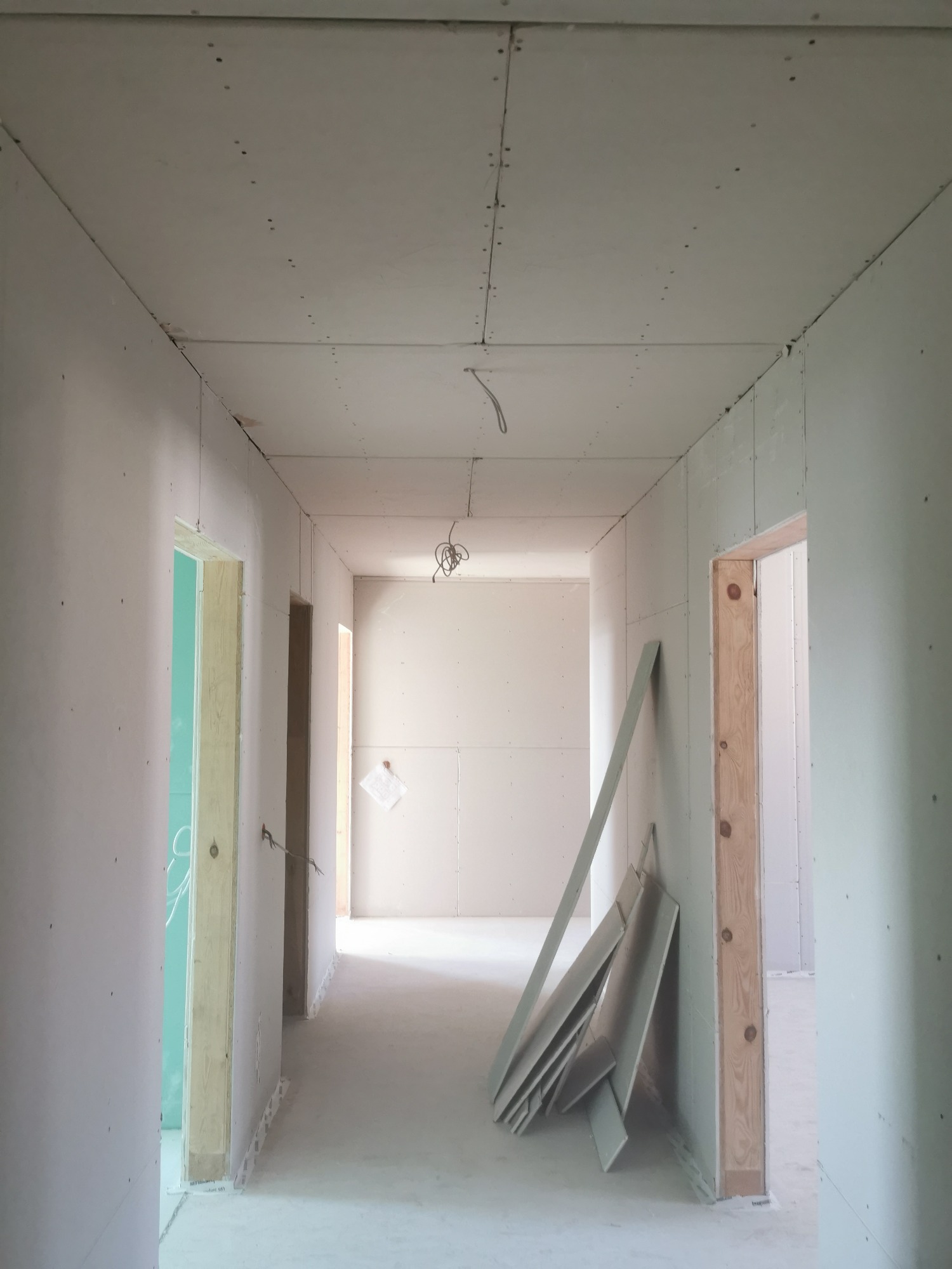 Korytarz w surowym stanie podczas prac wykończeniowych, ściany i sufit pokryte płytami gipsowo-kartonowymi, widoczne profile i przewody elektryczne, przygotowanie do dalszych prac.