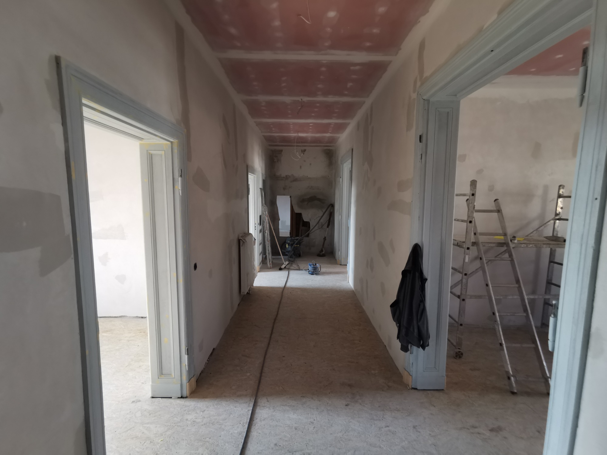 Korytarz w trakcie przygotowania do malowania: ściany po szpachlowaniu, sufit z płyt gipsowo-kartonowych, widoczne drabiny i narzędzia.