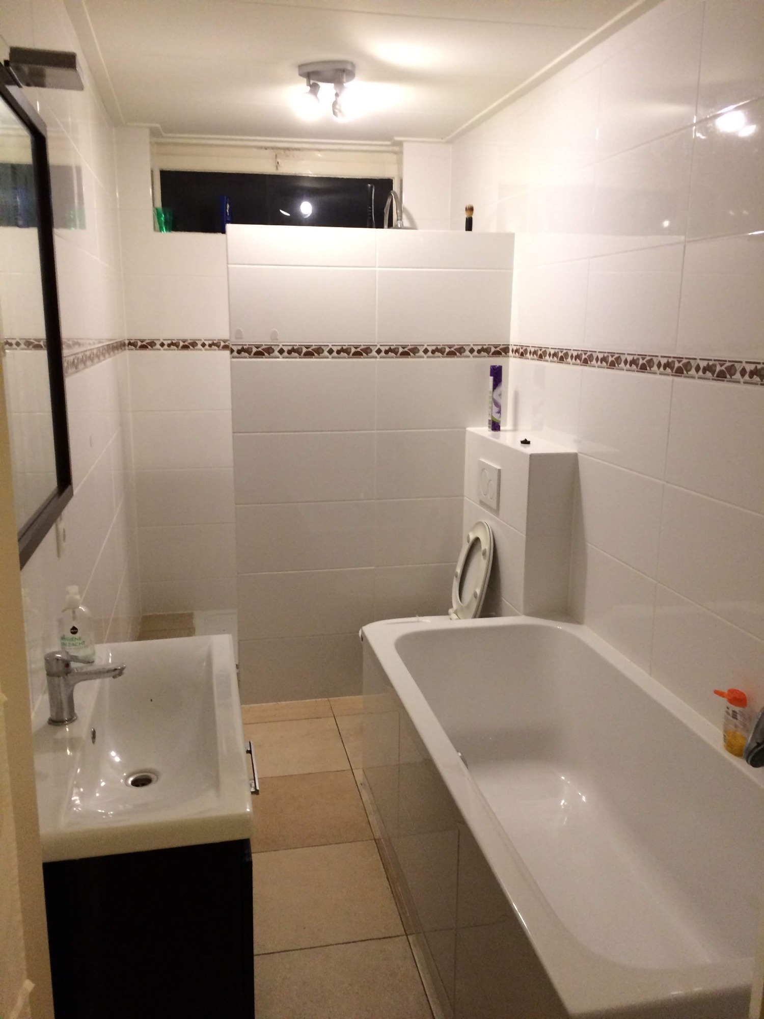 Wnętrze łazienki z białą wanną, umywalką z szafką w kolorze ciemnego brązu, toaletą ze spłuczką w zabudowie, wykończonej białymi płytkami z dekoracyjnym pasem na ścianach i brązową podłogą.