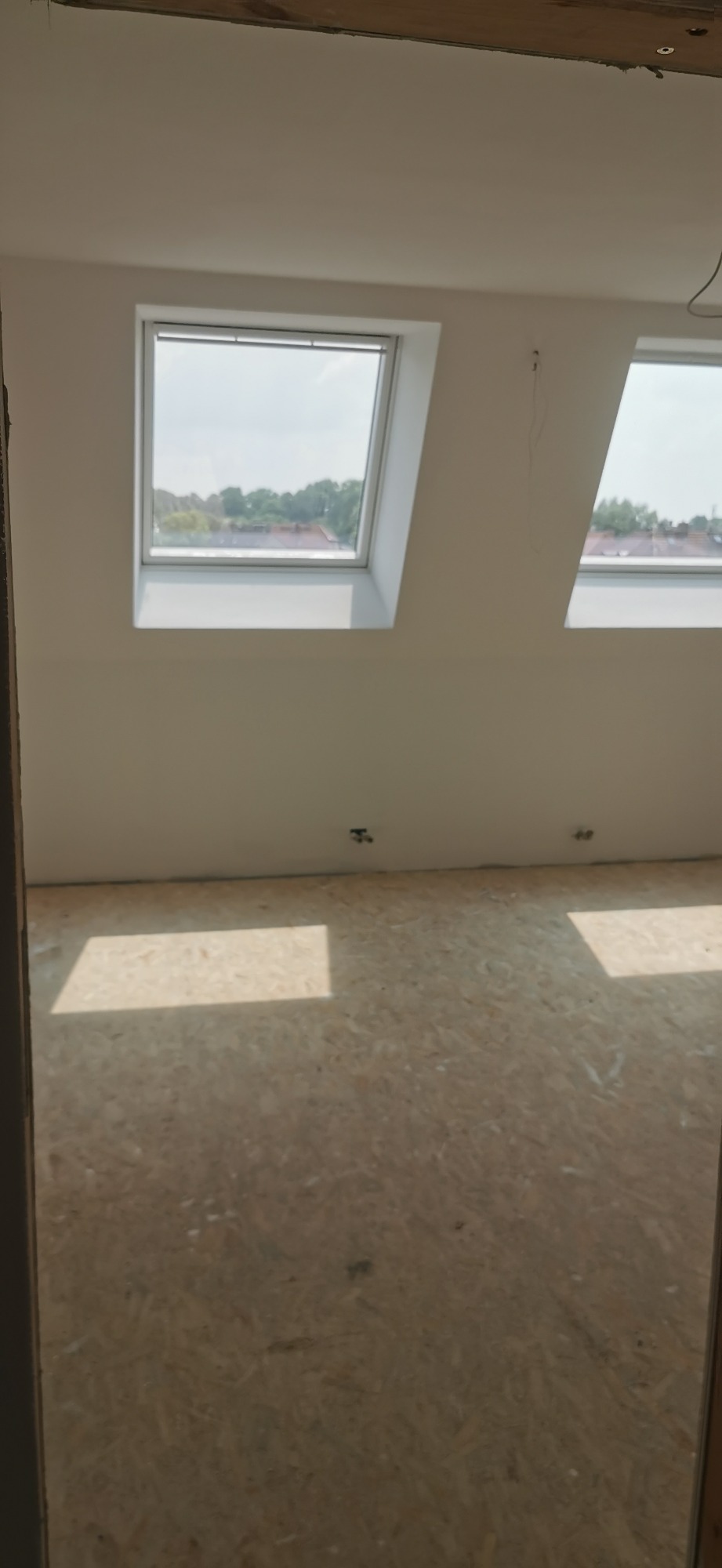 Poddasze w stanie deweloperskim z dwoma oknami dachowymi, surowe ściany pomalowane na biało, podłoga wyłożona płytami OSB, widok z niedokończonego wejścia.