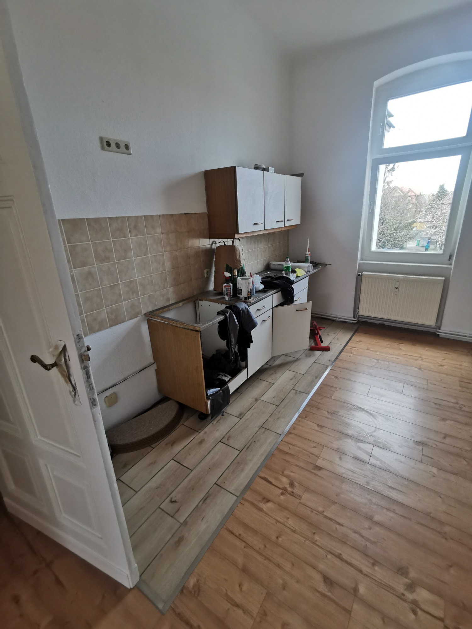Demontaż szafek kuchennych i płytek ściennych w trakcie prac przygotowawczych do remontu kuchni, widoczne odsłonięte gniazdko elektryczne, częściowo ułożona podłoga z paneli, okno z grzejnikiem.