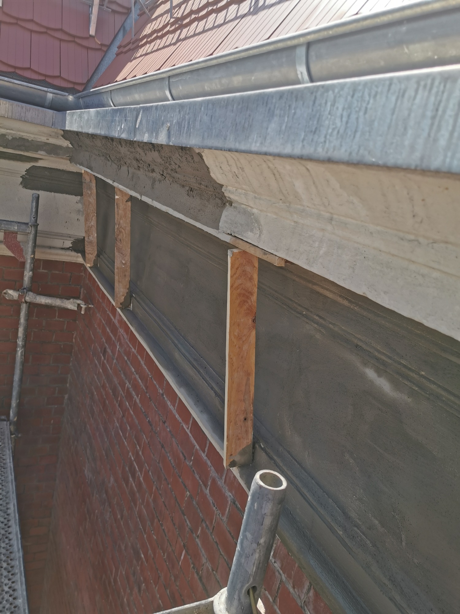 Fragment elewacji budynku z cegły w trakcie renowacji, widoczne szare tynki i drewniane podpory, rynna i fragment dachu w tle.