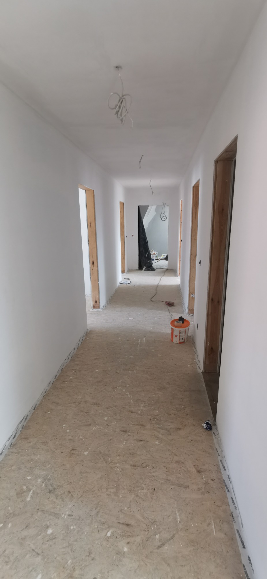 Korytarz w trakcie malowania na biało, widoczne gołe przewody oświetleniowe, surowa podłoga i otwarte ościeżnice drzwiowe.