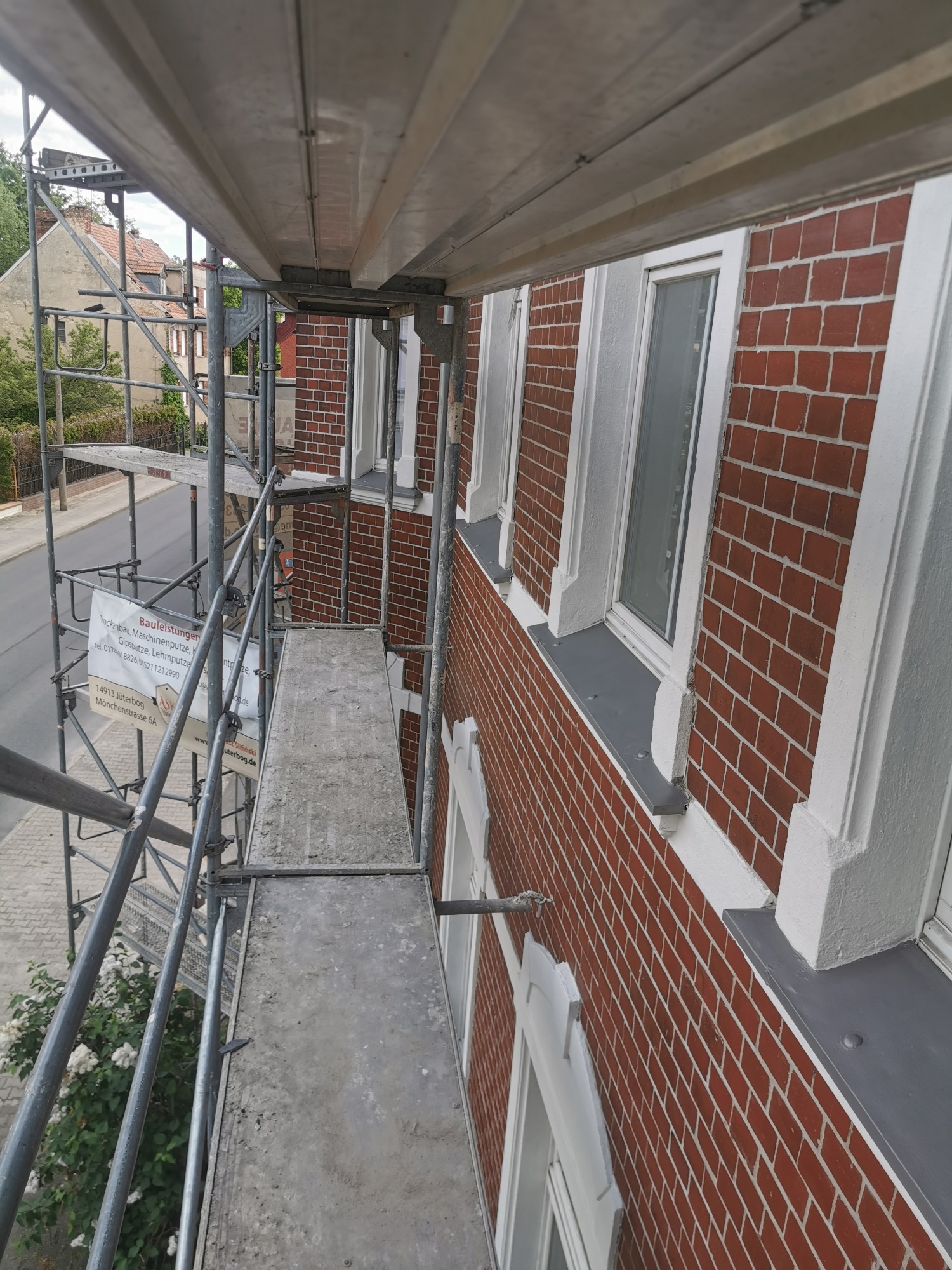 Ceglana elewacja budynku z białymi obramowaniami okien, widoczna z rusztowania podczas prac renowacyjnych.