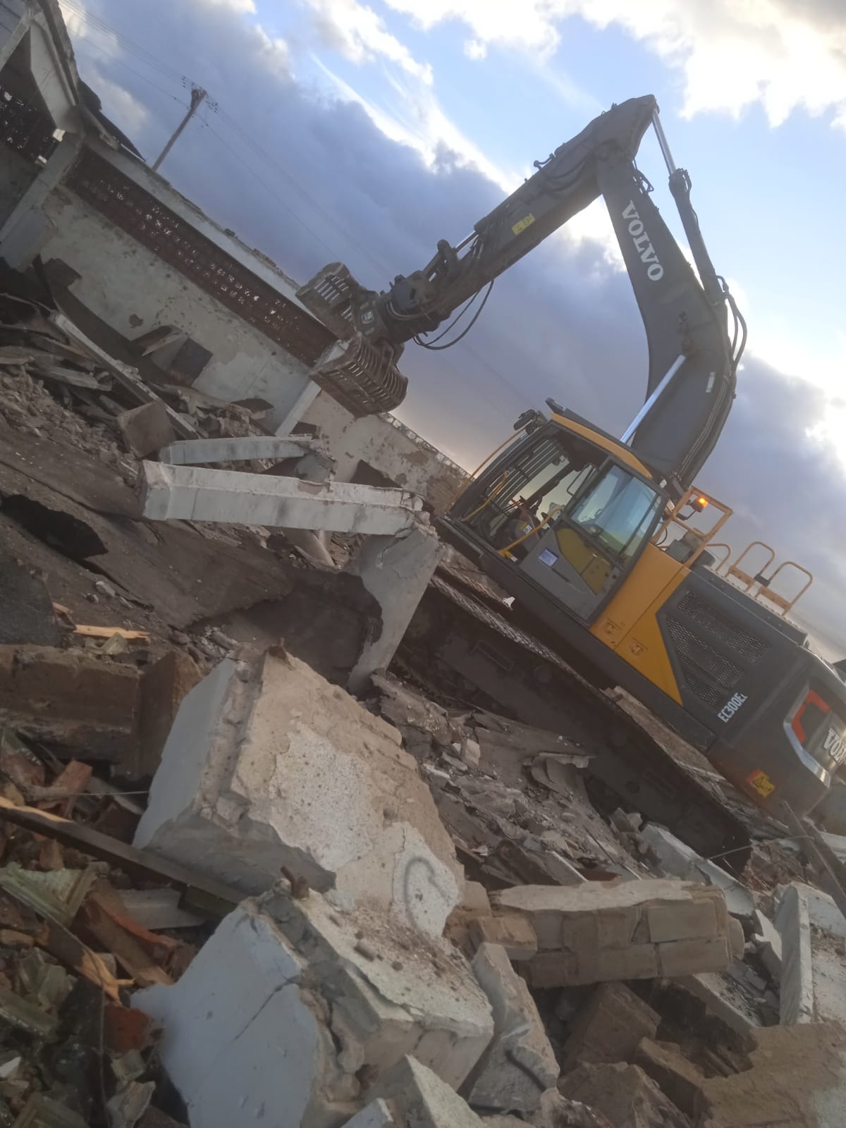 Żółto-czarna koparka Volvo EC300EL chwyta gruz na placu budowy podczas rozbiórki, widoczne fragmenty zburzonego budynku i pochmurne niebo.