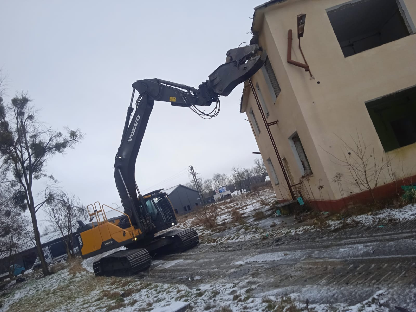 Żółto-czarna koparka Volvo z chwytakiem wyburza budynek z beżową elewacją, widoczne resztki śniegu na ziemi.