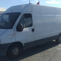 Biały, lekko zabrudzony bus dostawczy zaparkowany na brukowanym placu przed budynkiem magazynowym w słoneczny dzień.