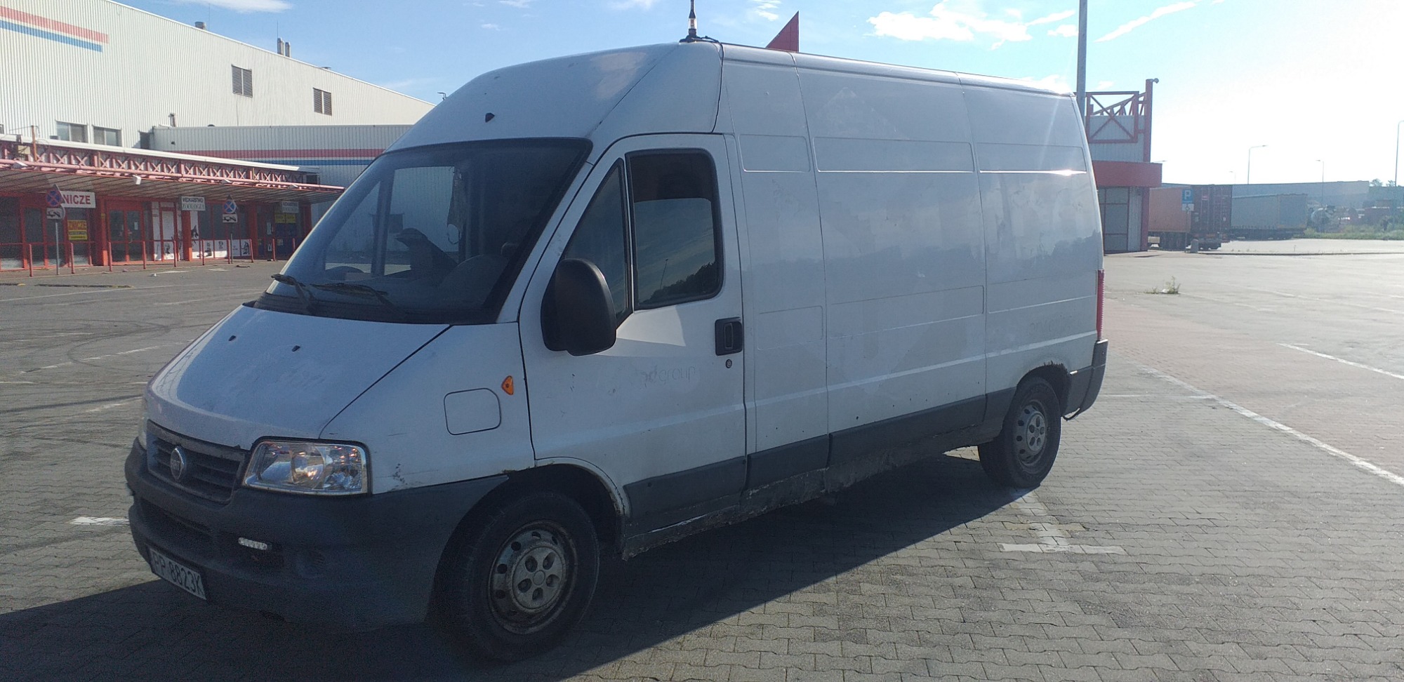 Biały, lekko zabrudzony bus dostawczy zaparkowany na brukowanym placu przed budynkiem magazynowym w słoneczny dzień.