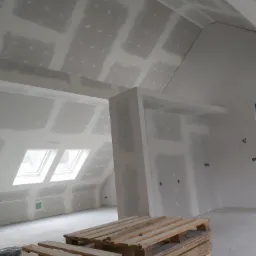 Poddasze po montażu płyt kartonowo-gipsowych, widoczne szpachlowane łączenia, okna dachowe, palety z płytami na podłodze.