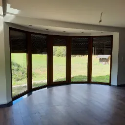 Jasne, puste wnętrze z drewnianą podłogą i panoramicznym oknem z brązowymi roletami, przez które widać zielony trawnik i skład drewna.