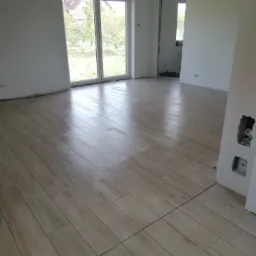 Jasne, drewnopodobne płytki ułożone na podłodze w nowym, jeszcze nieumeblowanym pomieszczeniu z dużym oknem i widokiem na ogród.