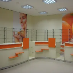 Puste wnętrze sklepu z ladami i regałami w kolorach kremowym i pomarańczowym, z grafiką na ścianie przedstawiającą pomarańcze i twarz kobiety z kremem, oświetlone przez panele LED w suficie.