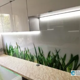 Nowoczesna kuchnia z białymi szafkami i blatem w odcieniach beżu, pomiędzy szafkami wiszącymi a blatem zamontowane szkło hartowane z nadrukiem motywu trawy, podświetlone od góry listwą LED.