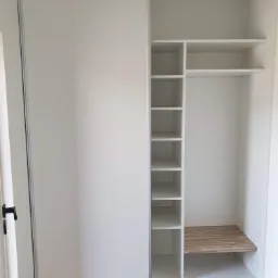 Wnętrze białej szafy wnękowej z przesuwnymi drzwiami, widoczne półki i drewniana wstawka na dolnej półce, oświetlenie sufitowe żarówką.