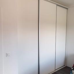 Expert Serwis bis sp z o o - Trzydrzwiowa szafa przesuwna w kolorze białym z szarymi obramowaniami, zamontowana w rogu pokoju z brązową podłogą i białymi ścianami, widoczny włącznik światła na ścianie.