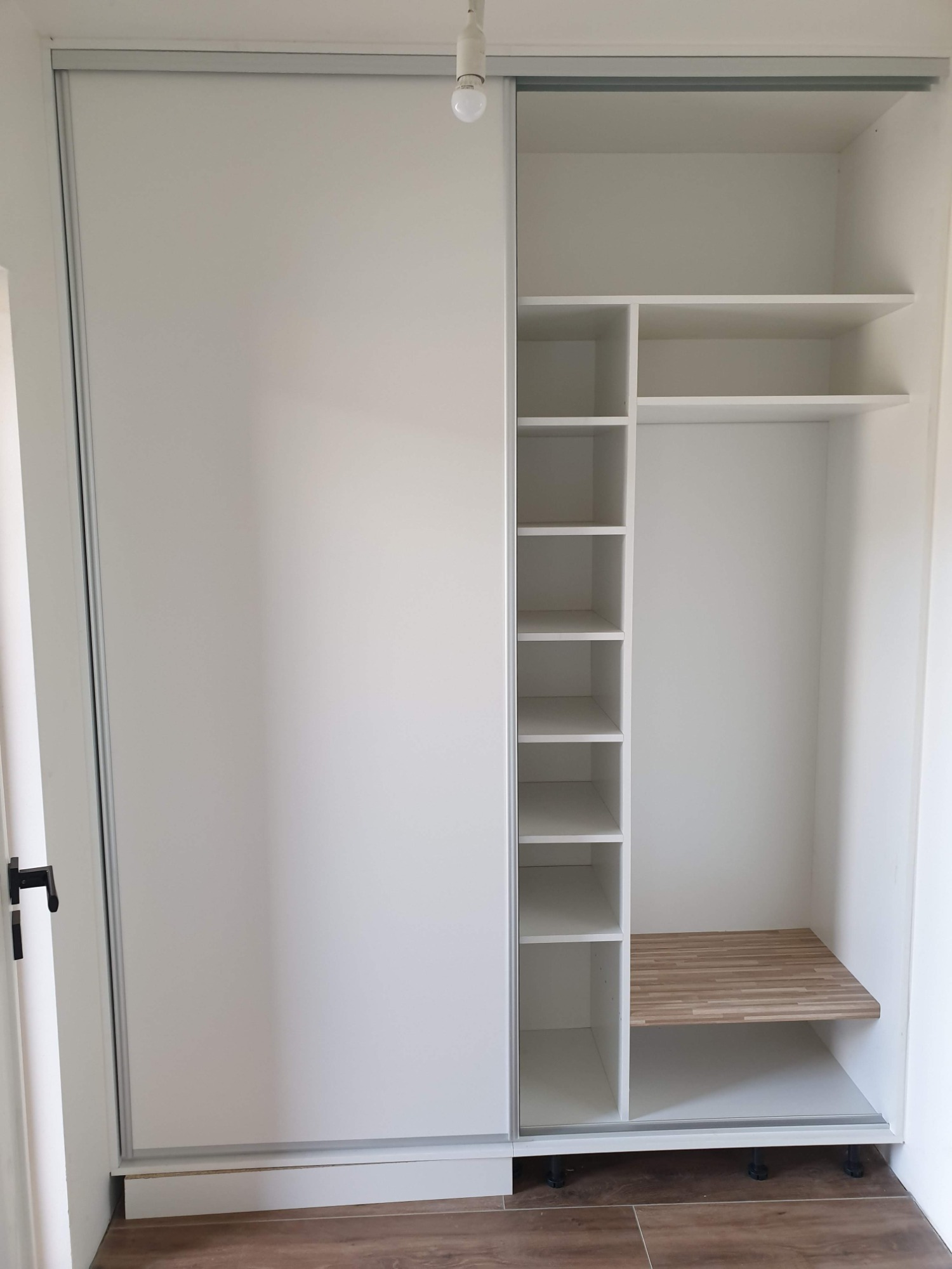 Wnętrze białej szafy wnękowej z przesuwnymi drzwiami, widoczne półki i drewniana wstawka na dolnej półce, oświetlenie sufitowe żarówką.