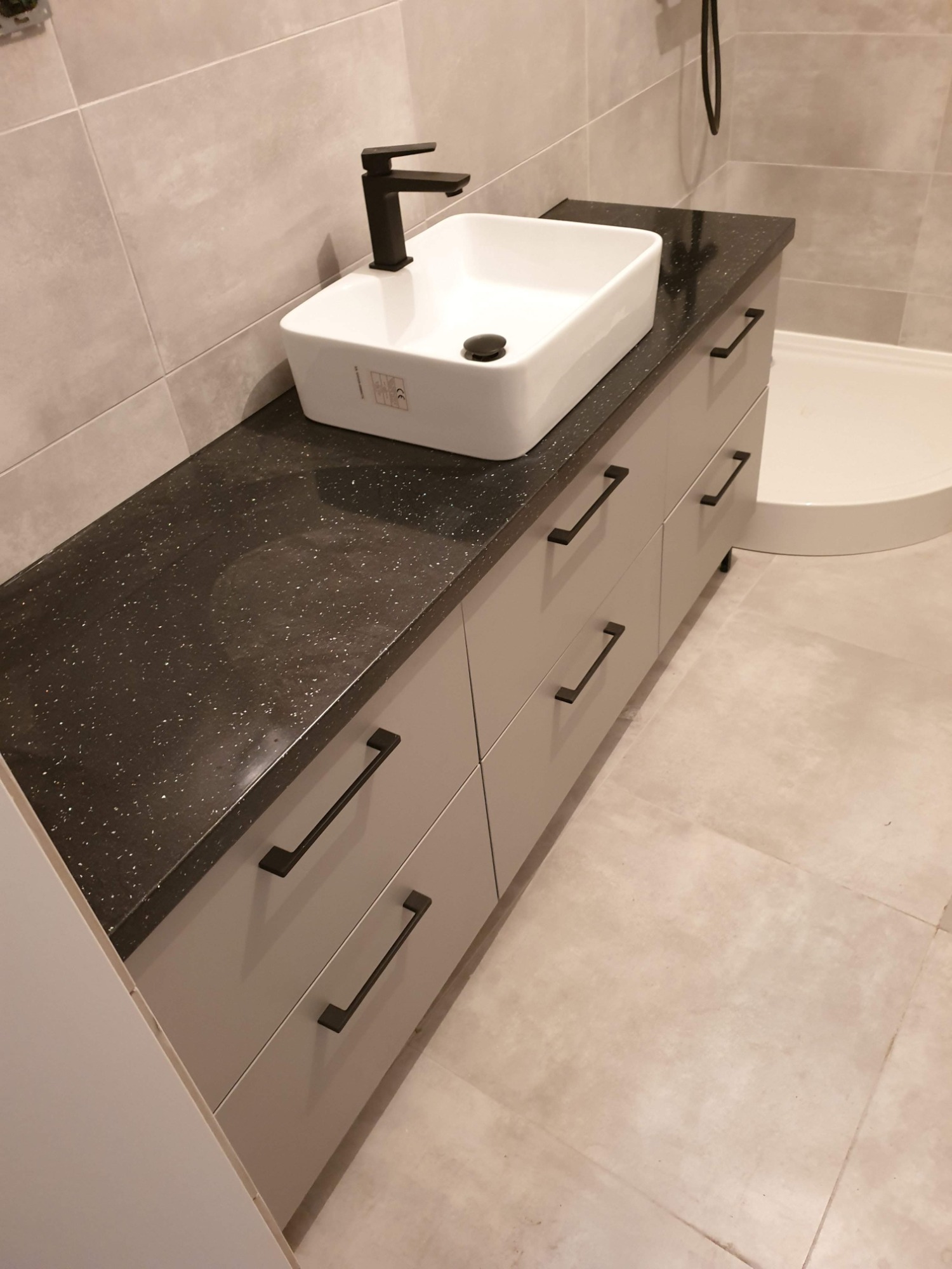 Łazienka z umywalką nablatową o kwadratowym kształcie i czarną armaturą, osadzona na szafce z szufladami w odcieniach szarości i blatem z ciemnego konglomeratu z jasnymi drobinkami, w tle widoczny...