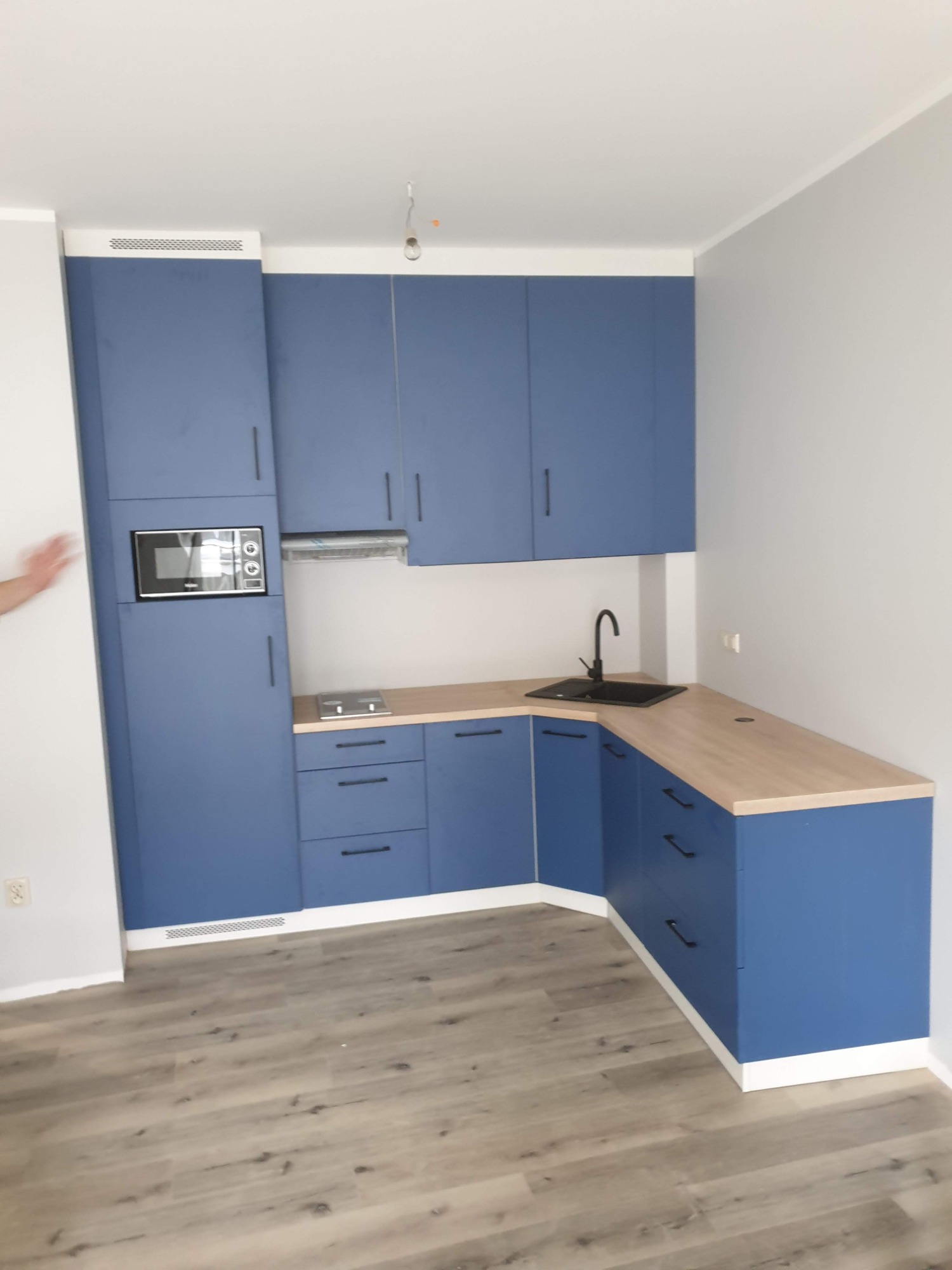 Niebieska zabudowa kuchenna z drewnianym blatem, czarnym zlewem i mikrofalówką w wysokiej szafce, widok na narożnik blatu i podłogę z paneli.
