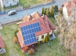Dom jednorodzinny z panelami słonecznymi na dachu, widok z góry z efektem miniatury, otoczony zielenią i zabudową mieszkalną.