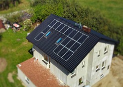 Widok z góry na dom jednorodzinny z zamontowanymi panelami fotowoltaicznymi na czarnym dachu blaszanym, dwa okna dachowe wkomponowane w instalację, komin z cegły. Otoczenie domu stanowi zieleń...