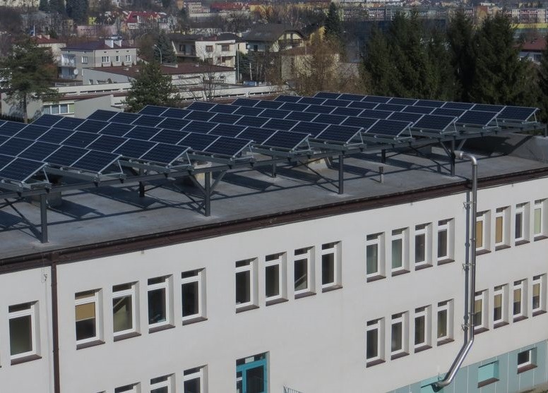 Rząd paneli słonecznych zamontowanych na metalowej konstrukcji nad płaskim dachem budynku, z widocznymi oknami i rurą spustową, na tle zabudowy miejskiej.