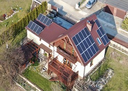 Dom jednorodzinny z brązowym dachem i panelami słonecznymi na dachu oraz na elewacji szczytowej, widok z góry.