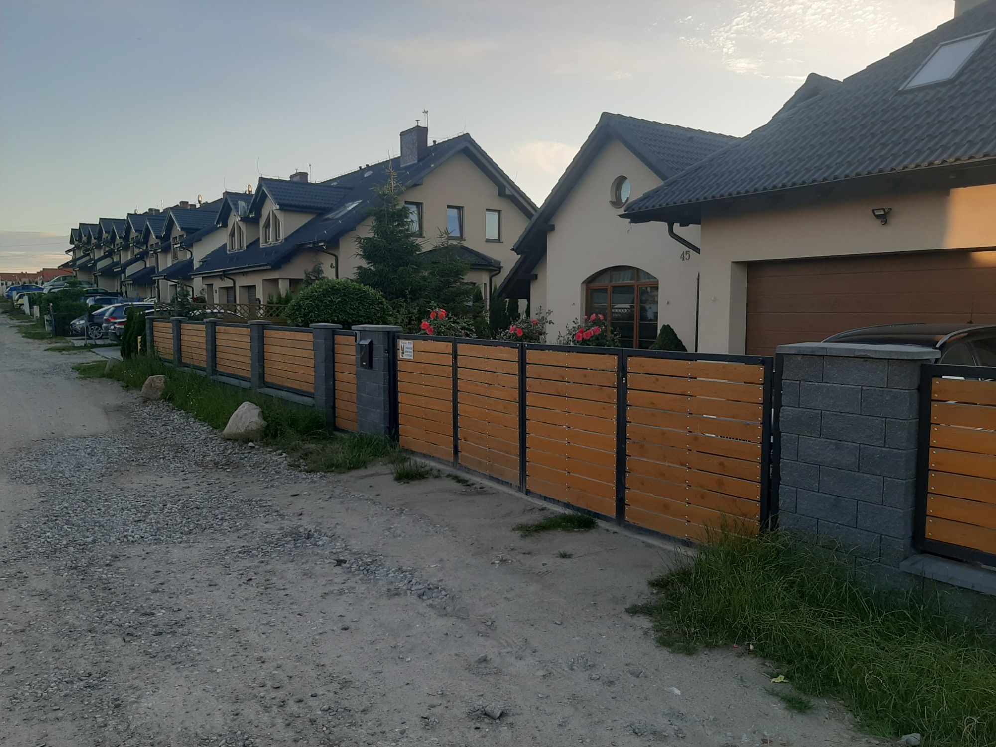 Długa, pozioma, drewniana ogrodzenie panelowe z szarymi, murowanymi słupkami, oddzielające szereg domów od żwirowej drogi.