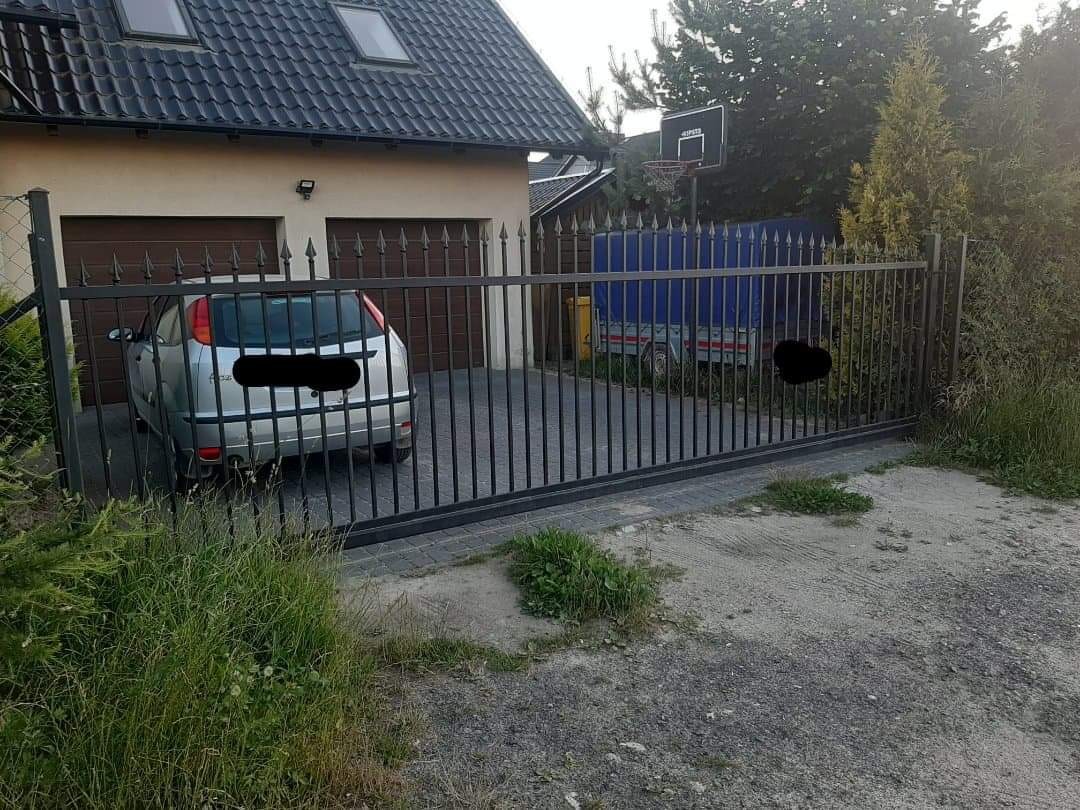 Czarna, metalowa brama przesuwna z ostrymi zakończeniami szprych, widoczna od strony podjazdu, z zaparkowanym samochodem i przyczepką w tle.