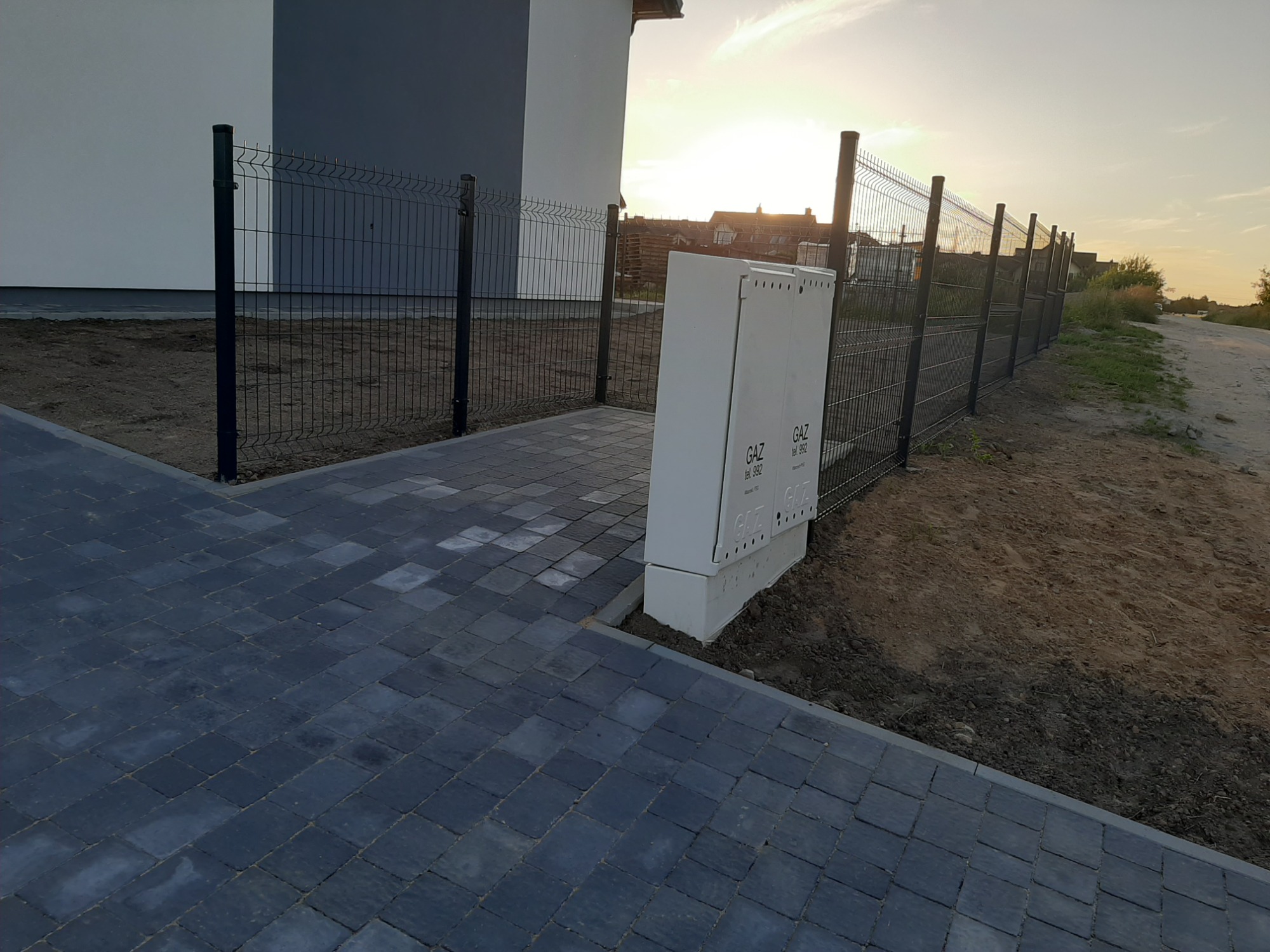 Nowoczesne ogrodzenie panelowe w kolorze grafitowym, zamontowane wzdłuż posesji z kostką brukową i skrzynką gazową, widok z perspektywy chodnika w słoneczny dzień.