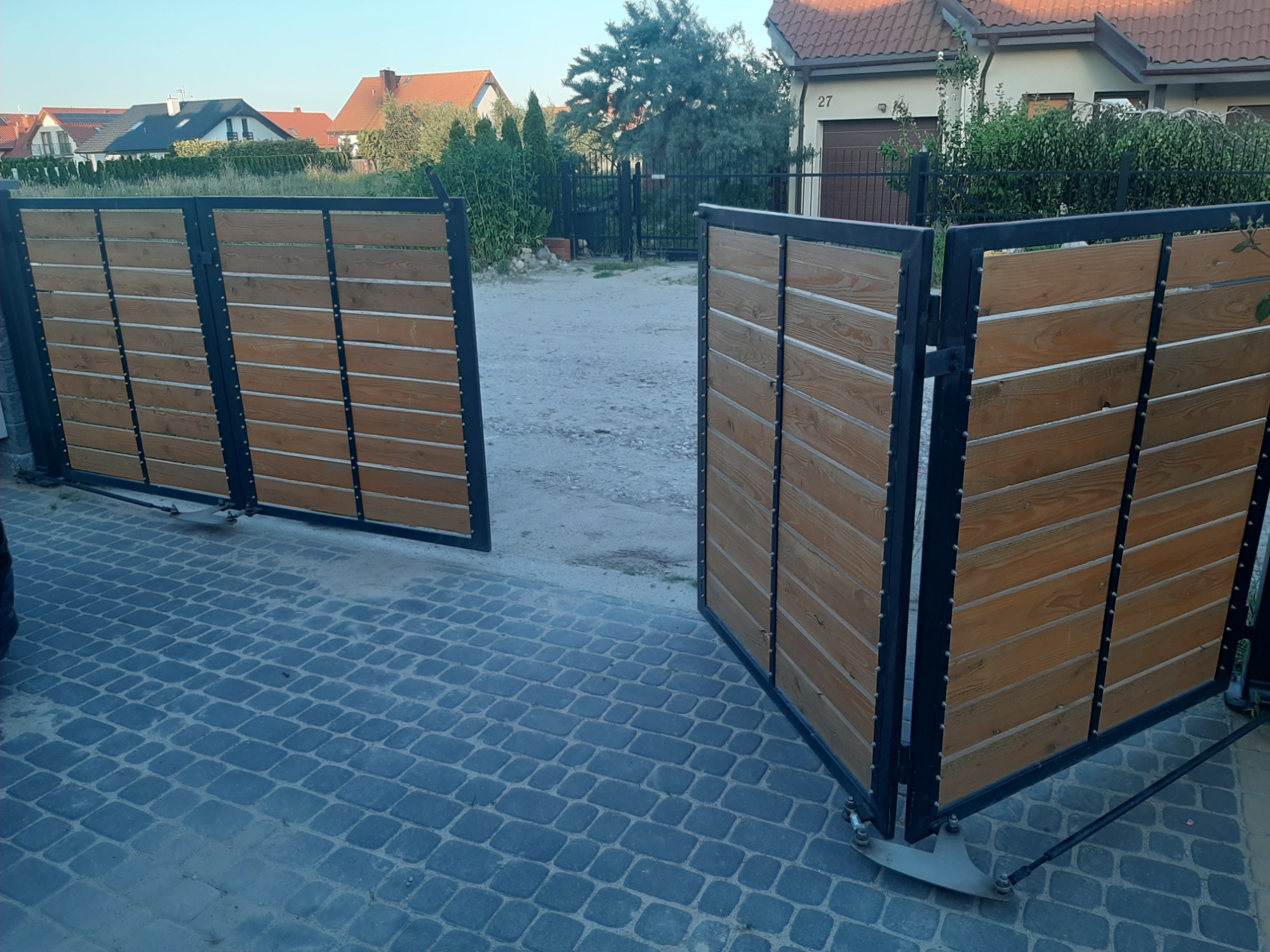 Otwarta, przesuwna brama z drewnianymi panelami i czarną, metalową ramą na posesji z kostki brukowej, widok z niskiej perspektywy.