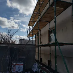 Elewacja budynku w trakcie prac, widoczne rusztowanie, ocieplenie styropianem w opakowaniach Arbet Eko Grafit, częściowo otynkowana ściana i błękitne niebo z chmurami.